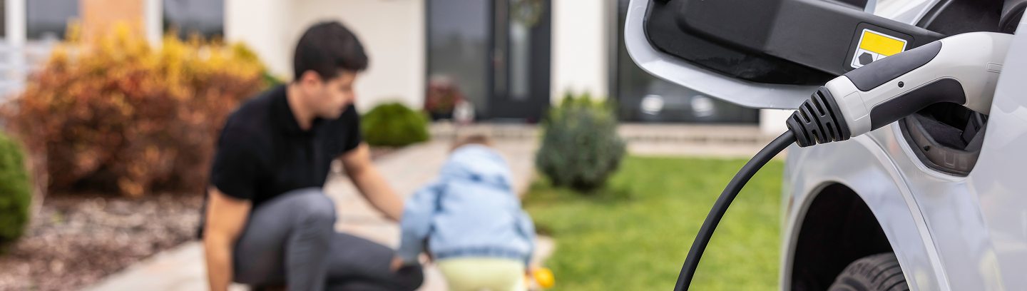 Image of a electric car charging in the background you see a father and child playing in their front yard.