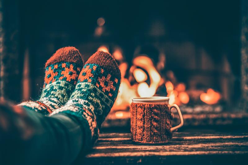 Feet in patterned socks resting by a fireplace with a mug in a knitted sleeve on a wooden surface