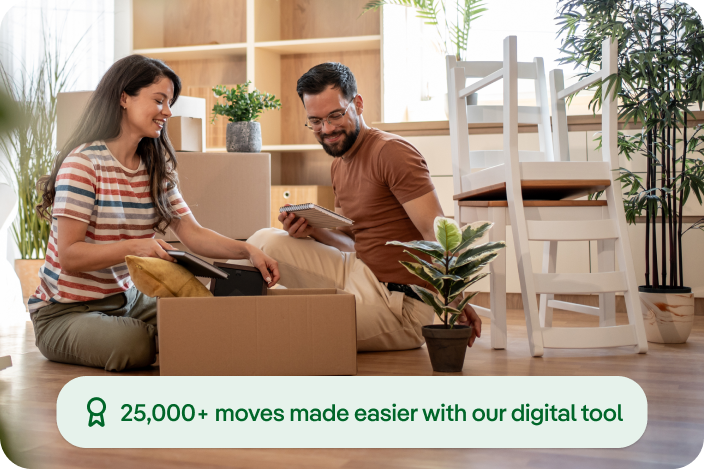 Two people sitting on the floor of a bright apartment with moving boxes, reviewing items on a tablet. On‑screen badge reads “25,000+ moves made easier with our digital tool.”